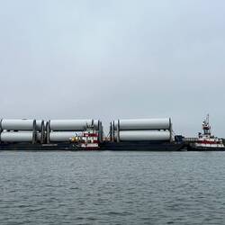 Two tugs moving a barge of windmill tower sections around.
