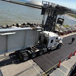 A massive structural item on a multi-wheel trailer is backed onto the ship