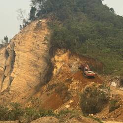 A solo man up high with pneumatic drill and digger both wrestle to tear away at the rock.